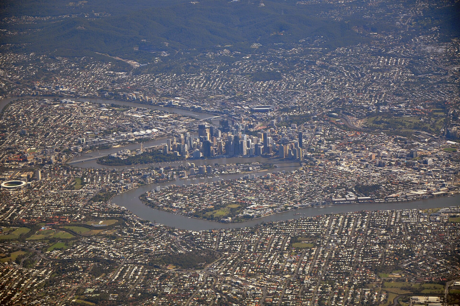 Aerial photograph of Brisbane looking across the winding Brisbane River, with the CBD's cluster of high-rise towers at centre, Suncorp Stadium and dense red-roofed suburbs spreading out to forested hills on the horizon.